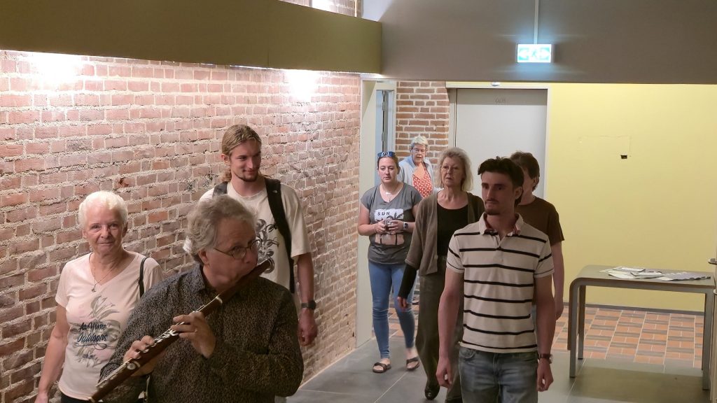 Opening muzieklokalen Cultuurschuur. Groep mensen volgt een meneer die op een dwarsfluit speelt door het nieuwe muziekcentrum