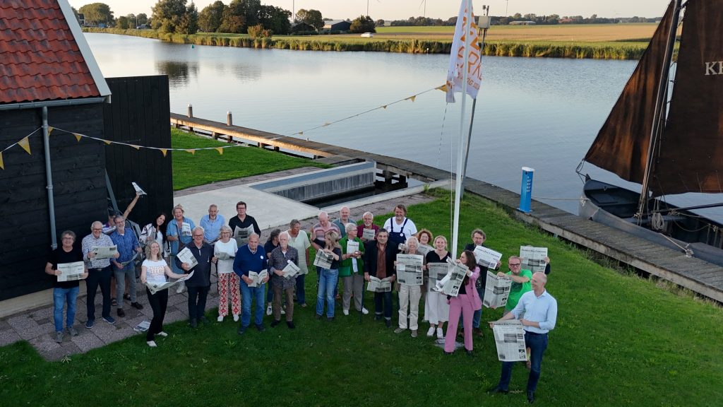 Groepsfoto van boven van alle aanwezigen met elk een krant in hun handen. Rechts het water en de jol KH44
