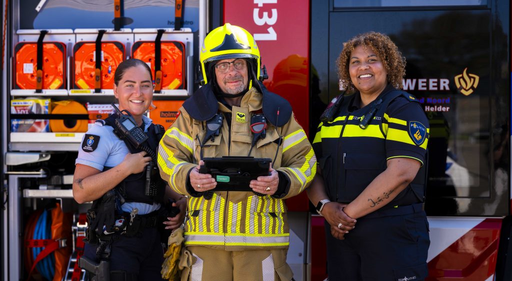 112 Hulpverleningsdag in Den Helder. Politie, brandweer en marechaussee staan naast elkaar in uniform en kijken de camera in. Op de achtergrond een brandweer en politieauto.