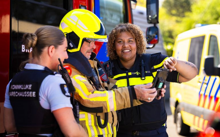 112 Hulpverleningsdag in Den Helder. Politie, brandweer en marechaussee staan naast elkaar in uniform en leggen iets uit. Op de achtergrond een brandweer en politieauto.
