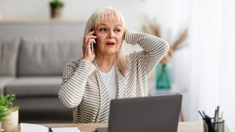 Mevrouw wordt telefonisch gescamd en zit achter laptop aan een bureau aan de telefoon en kijkt verschrikt.
