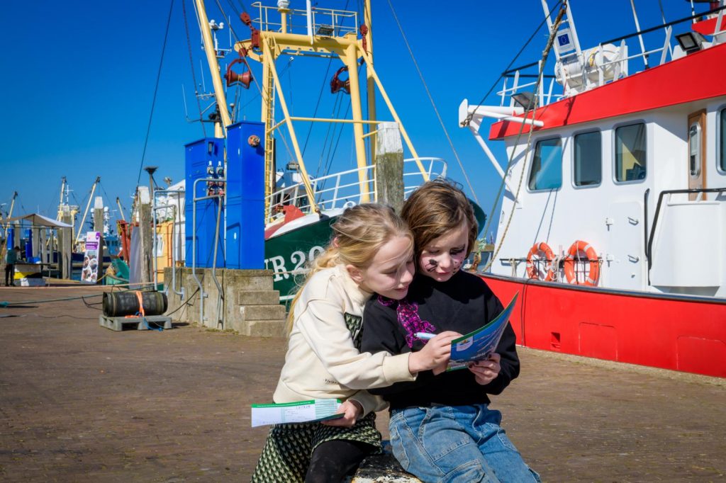 Kom Binnen Bij Bedrijven Dagen. Twee meisjes lezen een flyer in Den Oever voor wat vissersboten