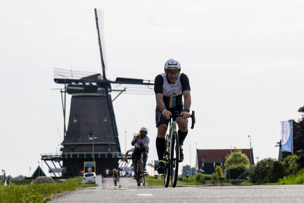 Wielrenners passeren een kruispunt in een dorp, terwijl toeschouwers langs de kant staan en applaudisseren. Op de achtergrond staan een karakteristiek huis en groen landschap.