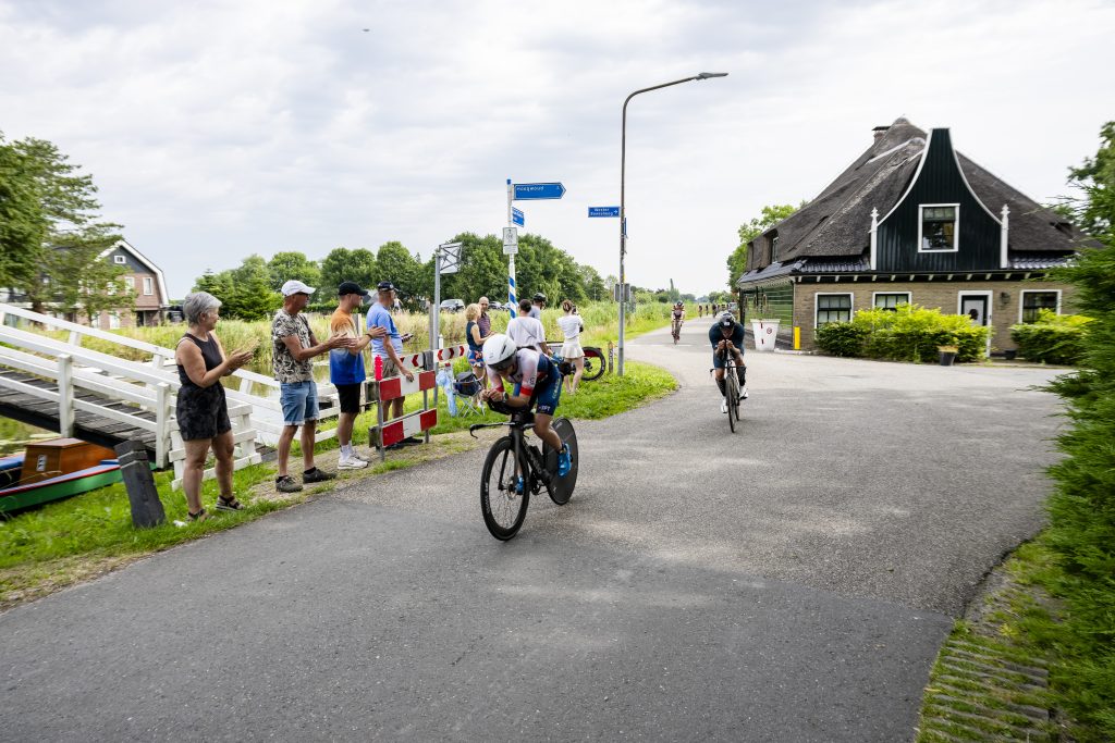 Wielrenner fietst in voorovergebogen houding op een rechte weg, met een traditionele windmolen op de achtergrond. Andere deelnemers volgen op afstand langs een landelijk parcours.