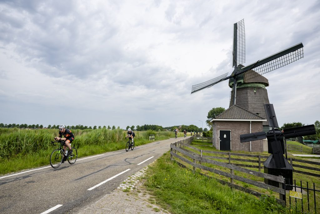 Groep wielrenners fietst over een landelijke weg langs een traditionele windmolen met rieten kap. De weg loopt door groen polderlandschap met riet en weilanden onder een bewolkte lucht.