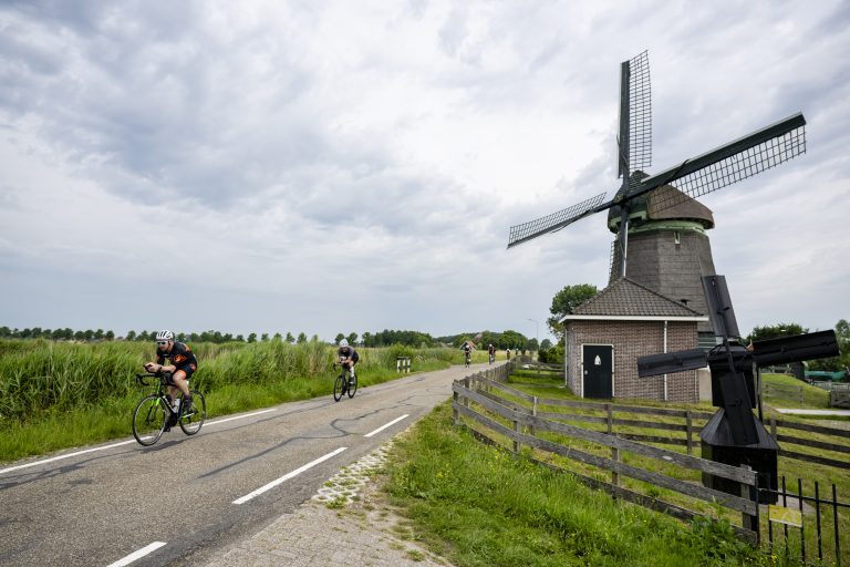 Groep wielrenners fietst over een landelijke weg langs een traditionele windmolen met rieten kap. De weg loopt door groen polderlandschap met riet en weilanden onder een bewolkte lucht.