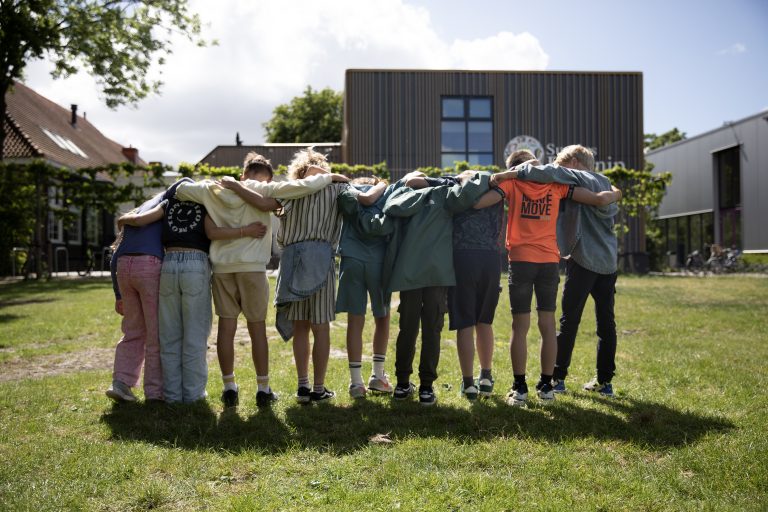Foto van een groep kinderen die met hun armen over elkaars schouder in een rij met hun rug naar de camera voor een school staan op een grasveld