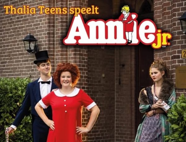 Promotiefoto van de musical Annie Jr.. Op de voorgrond staan drie acteurs in kostuum voor een bakstenen gebouw. Bovenaan staat de tekst: “Thalia Teens speelt Annie Jr.”. In het midden staat een acteur in een rode jurk, geflankeerd door twee andere acteurs in theaterkleding die passen bij de musical.