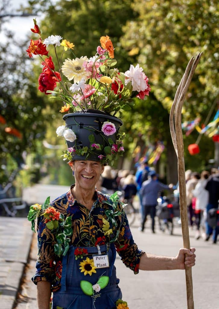Peter Plant, meneer met overall aan waarop bloemen vastzitten. Op zijn hoofd een emmer met bloemen. In zijn hand een houten stok.