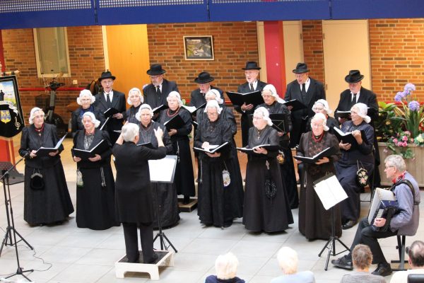 Koor in traditionele Wieringer klederdracht treedt op in een binnenruimte. De zangers en zangeressen staan in meerdere rijen met muziekmappen in de hand, terwijl een dirigent voor het koor staat. Rechts begeleidt een accordeonist het optreden. Op de achtergrond zijn bakstenen muren en bloemen te zien.