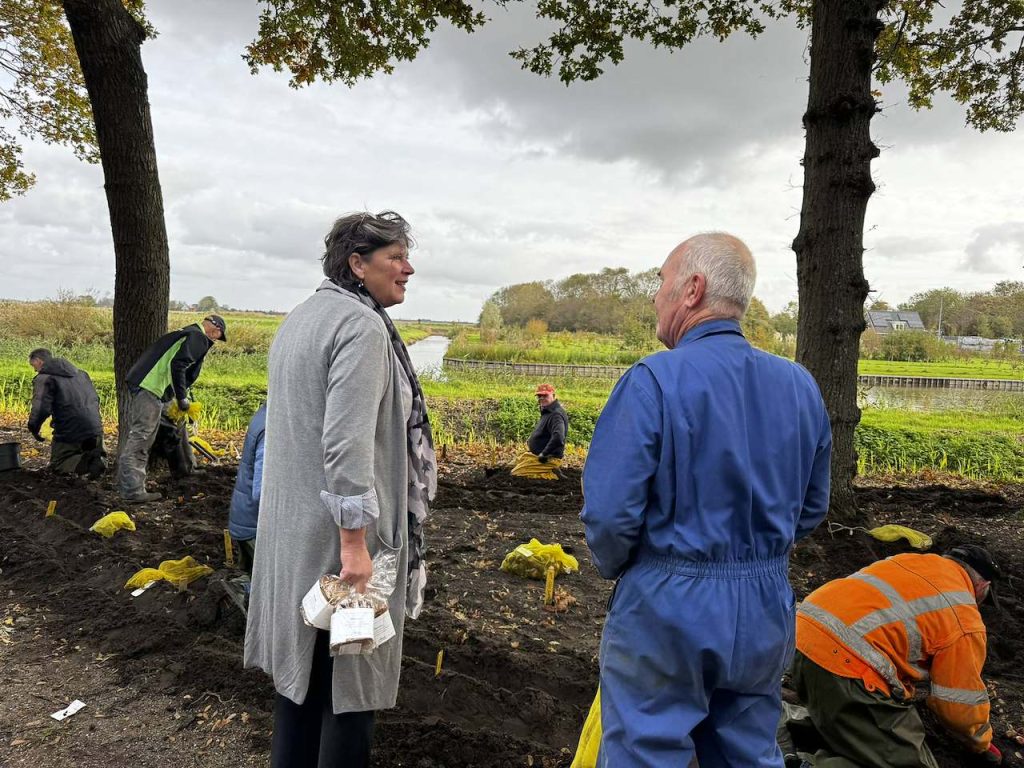 Overzicht van de Poldertuin in Anna Paulowna, waar vrijwilligers duizenden bloembollen hebben geplant. In de donkere aarde tussen de bomen staan veel gele bordjes die de verschillende plantplekken aangeven. Op de grond liggen nog enkele gele zakken met bloembollen. Burgemeester Rian van Dam kwam langs namens de gemeenteraad met koeken voor de vrijwilligers.