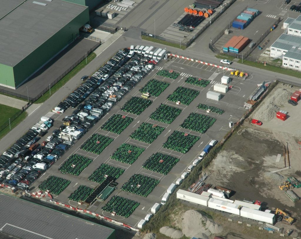 Luchtfoto van een bedrijventerrein met duizenden groen-zwarte GO Sharing-deelscooters die in nette vakken geparkeerd staan op een groot parkeerterrein. Naast de scooters staan rijen geparkeerde auto's, bedrijfsgebouwen en enkele containers en vrachtwagens.
