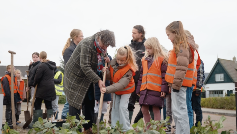 Een groep kinderen en burgemeester Rain van Dam plant samen groen in een border. Een kind gebruikt samen met een volwassene een schep, terwijl andere kinderen toekijken.