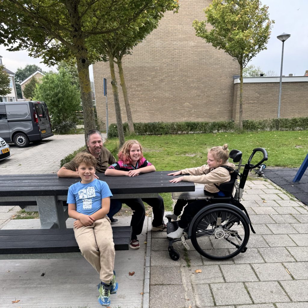 Roger Voetel deed als buurtbewoner een oproep deed voor het aanpassen van de speeltuin voor dochter Yamilia. Een groep mensen zit en staat rond een picknicktafel in een speeltuin of openbare ontmoetingsplek in een woonwijk. Aan de tafel zitten twee kinderen en een volwassene, terwijl een derde kind in een rolstoel aan de tafel plaatsneemt. Op de achtergrond zijn bomen, gras, een speeltoestel en woningen zichtbaar. De foto laat een toegankelijke ontmoetingsplek zien waar ook ruimte is voor een rolstoel.