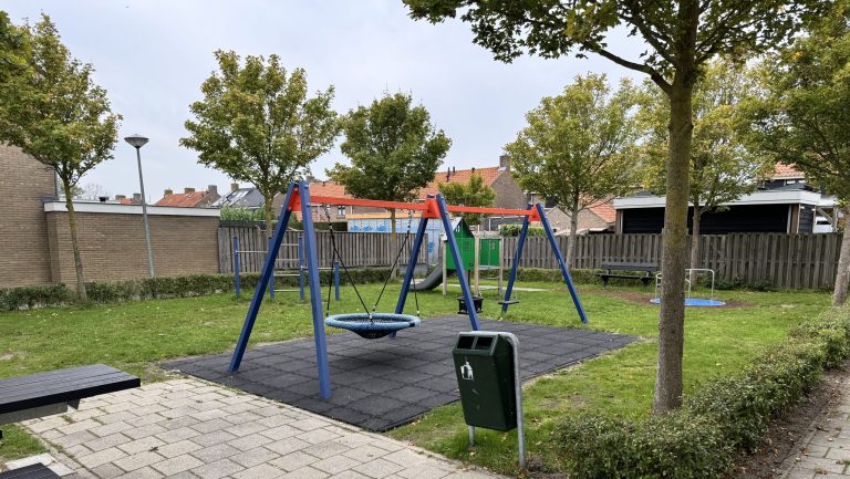 Overzicht van een speeltuin met een nestschommel, draaiplateau, glijbaan en zitgelegenheid. De speeltoestellen staan op een ruim grasveld tussen bomen en woningen.