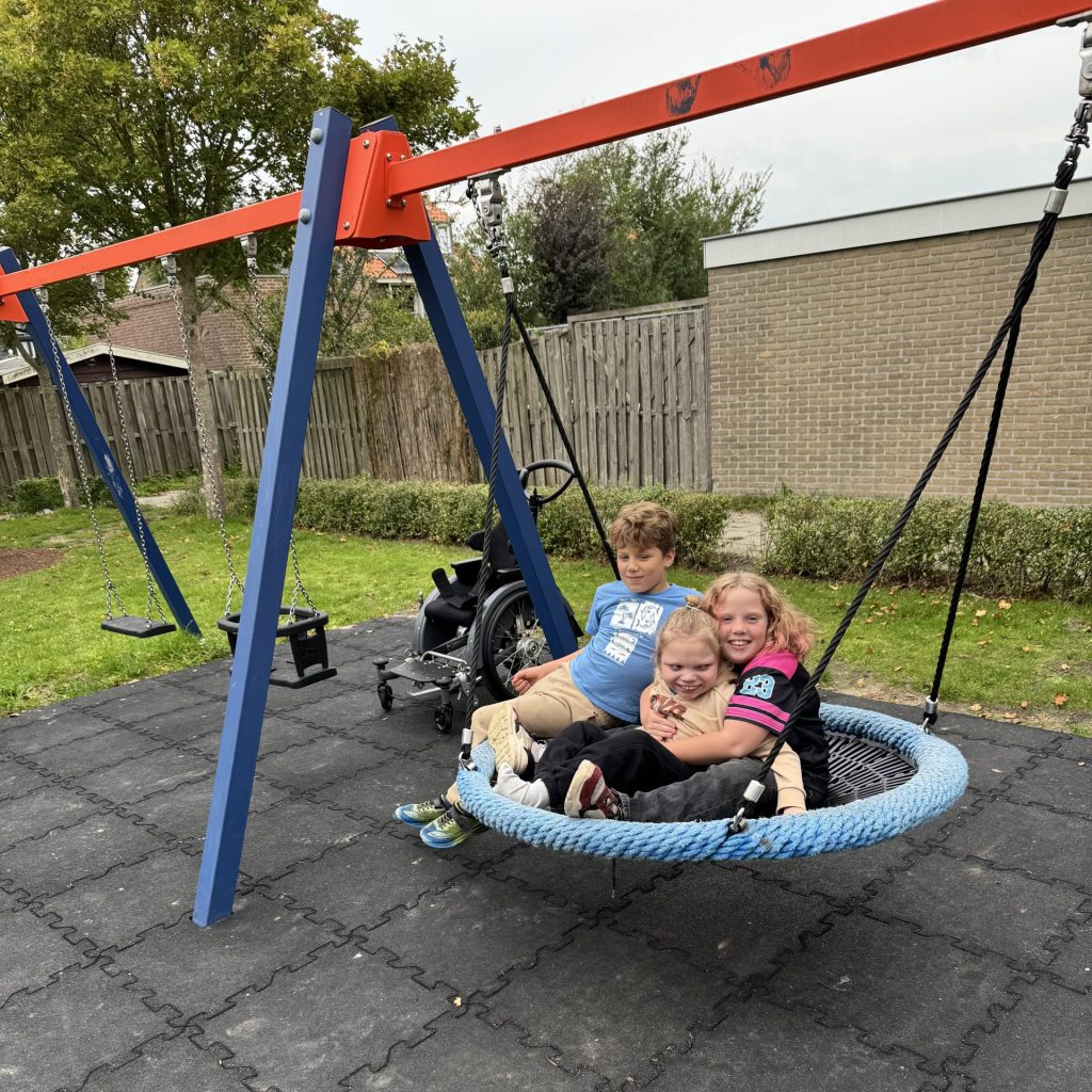 Drie kinderen maken gebruik van een grote nestschommel in een speeltuin. Naast de schommel staat een rolstoel. De speelplek heeft een zachte rubberen ondergrond en wordt omringd door gras, bomen en een houten erfafscheiding.
