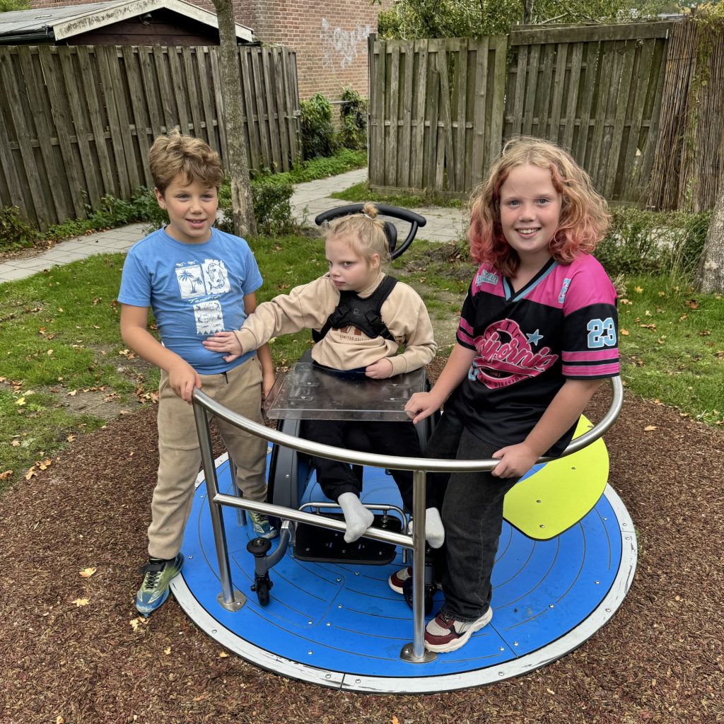 Drie kinderen gebruiken samen een toegankelijk draaiplateau. Een kind zit met een rolstoel op het speeltoestel, terwijl twee andere kinderen ernaast staan. Rondom het toestel ligt een valdempende ondergrond van houtsnippers.
