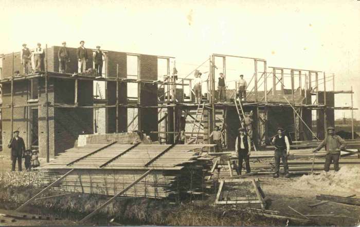 Historische zwart-witfoto van de bouw van een bollenschuur. Het gebouw bevindt zich in een vroeg stadium van de bouw, met zichtbare bakstenen muren, houten constructies en steigers. Op en rond de steigers staan meerdere bouwvakkers. Op de voorgrond liggen stapels bouwmaterialen, waaronder houten platen en balken. De omgeving is open en landelijk, met een sloot langs de bouwplaats. De foto geeft een beeld van de aanleg van een bedrijfsgebouw in de bollenteelt.