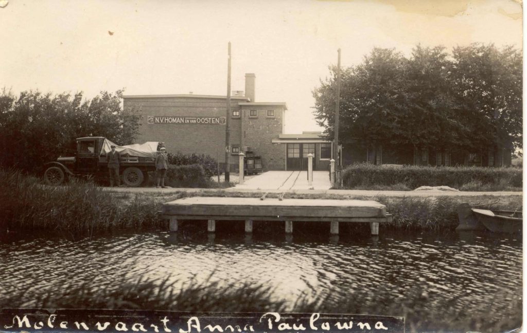 Historische zwart-witfoto van een bedrijfsgebouw aan het water in Anna Paulowna. Op de gevel staat de tekst “N.V. Homan en Van Oosten”. Op de voorgrond ligt een houten aanlegsteiger langs een vaart. Links staat een vrachtwagen met afgedekte lading en twee personen naast het voertuig. Het bedrijfsterrein is bereikbaar via een oprit tussen enkele palen. Rondom het gebouw staan bomen en struiken. Onderaan de foto staat handgeschreven: “Molenvaart Anna Paulowna”.