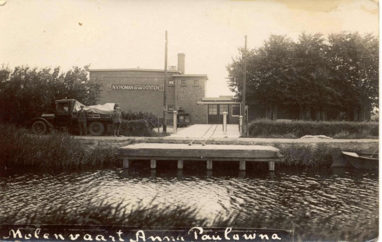 Historische zwart-witfoto van een bedrijfsgebouw aan het water in Anna Paulowna. Op de gevel staat de tekst “N.V. Homan en Van Oosten”. Op de voorgrond ligt een houten aanlegsteiger langs een vaart. Links staat een vrachtwagen met afgedekte lading en twee personen naast het voertuig. Het bedrijfsterrein is bereikbaar via een oprit tussen enkele palen. Rondom het gebouw staan bomen en struiken. Onderaan de foto staat handgeschreven: “Molenvaart Anna Paulowna”.